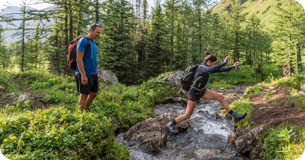 To fotturister som en om gangen hopper over en liten bekk midt i en skog